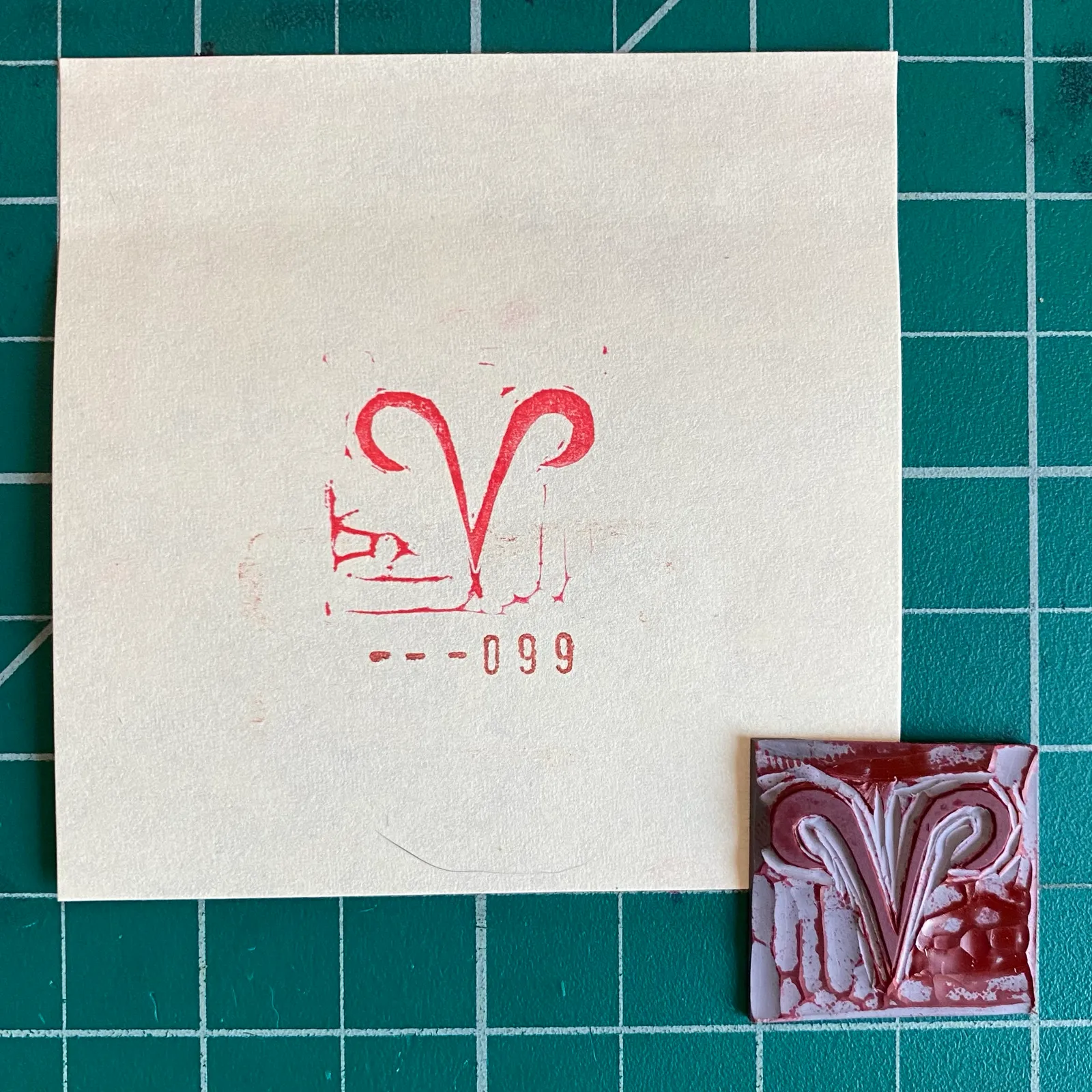 A cream square rests on a green grid, marked with red ink forming a stylized V and numbers; a small, textured square of deep red and white abstract patterns sits below it.