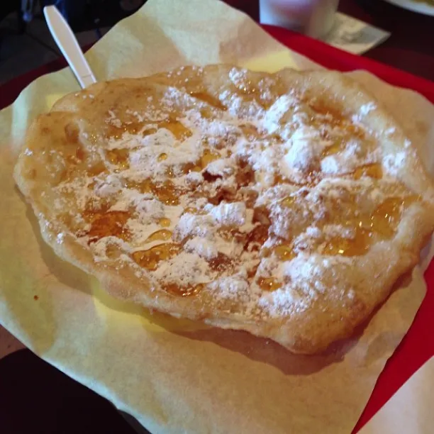 A golden-brown, flaky pastry with visible caramelized filling is centered on light yellow paper, dusted heavily with white powdered sugar, set against a red background.