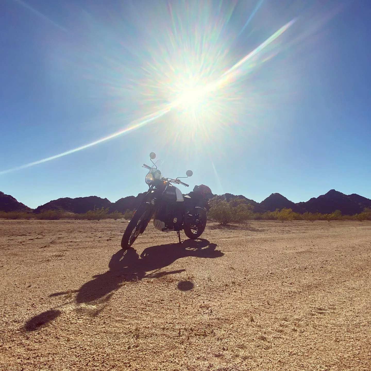 A motorcycle rests on granular tan ground, silhouetted against a vast desert expanse under a brilliant blue sky dominated by the sun's flare and long shadows.