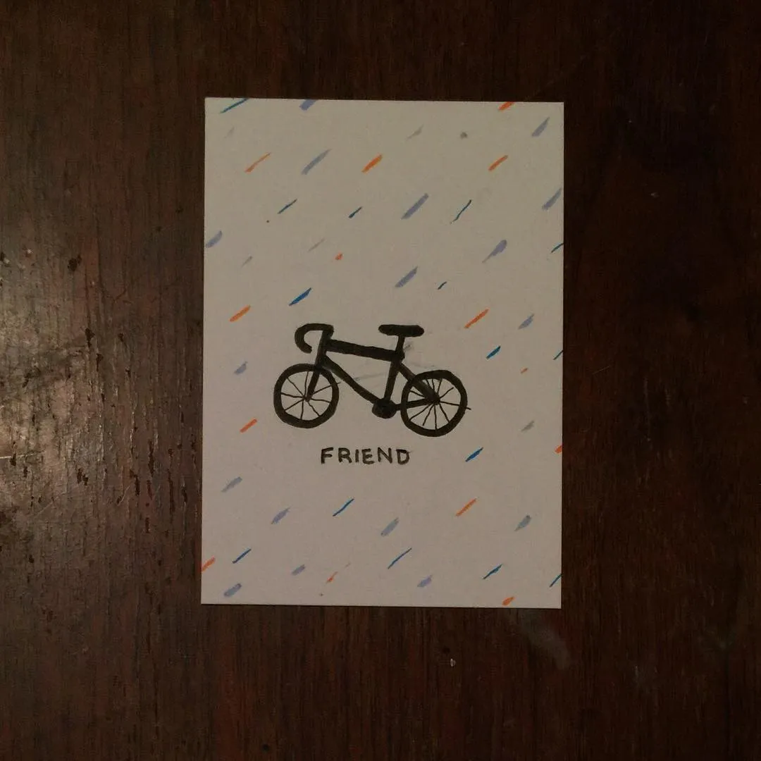 The card features a black line drawing of a bicycle and the word FRIEND, set against a background of scattered colored dashes in orange, blue, and red.