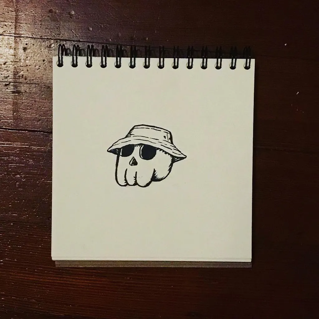 A black line drawing of a skull wearing sunglasses and a straw hat is centered on a small, spiral-bound notepad resting on a dark wooden surface.