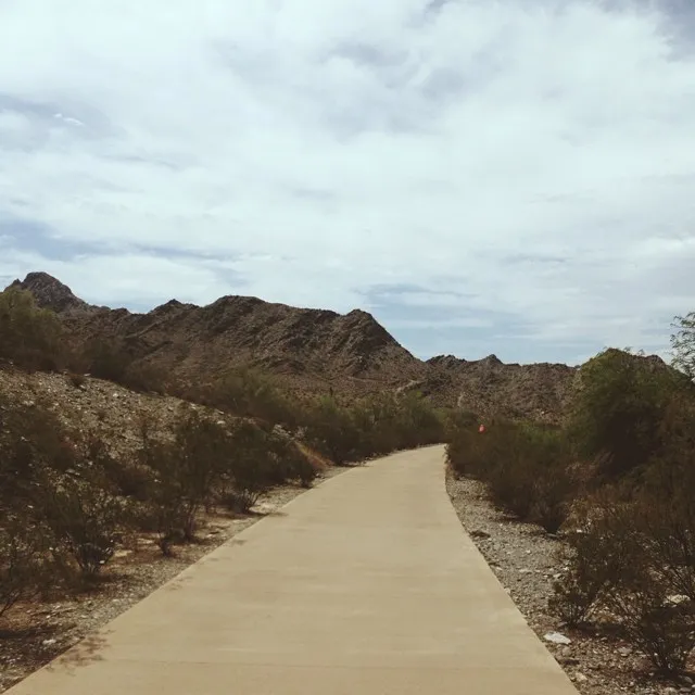 A smooth, curving path leads through rugged, brown hills covered in dark scrub brush, set beneath a vast, overcast sky.