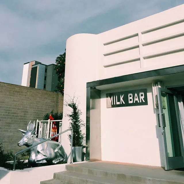 A stark white building facade with horizontal bands and a sign reading MILK BAR stands next to a metallic sculpture of a cow and concrete steps under a pale sky.