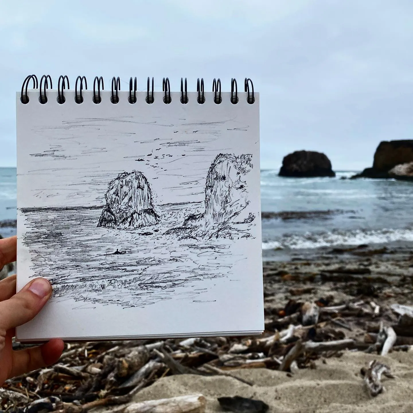 A black ink sketch in a spiral notebook depicts three large sea stacks in choppy water, with a row of drawn curved shapes positioned above the scene.