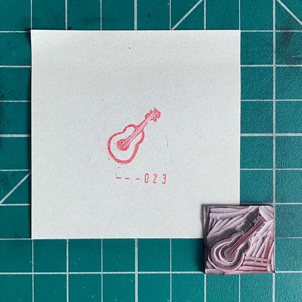 On a teal grid, a white paper displays a red outline drawing of a guitar and the numbers 223, next to a small, textured square with layered pink and gray forms.