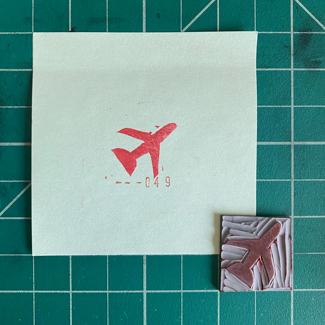 A pale square on a green grid holds a red airplane graphic and the numbers -049, positioned above a small, textured square of pink and gray folds.
