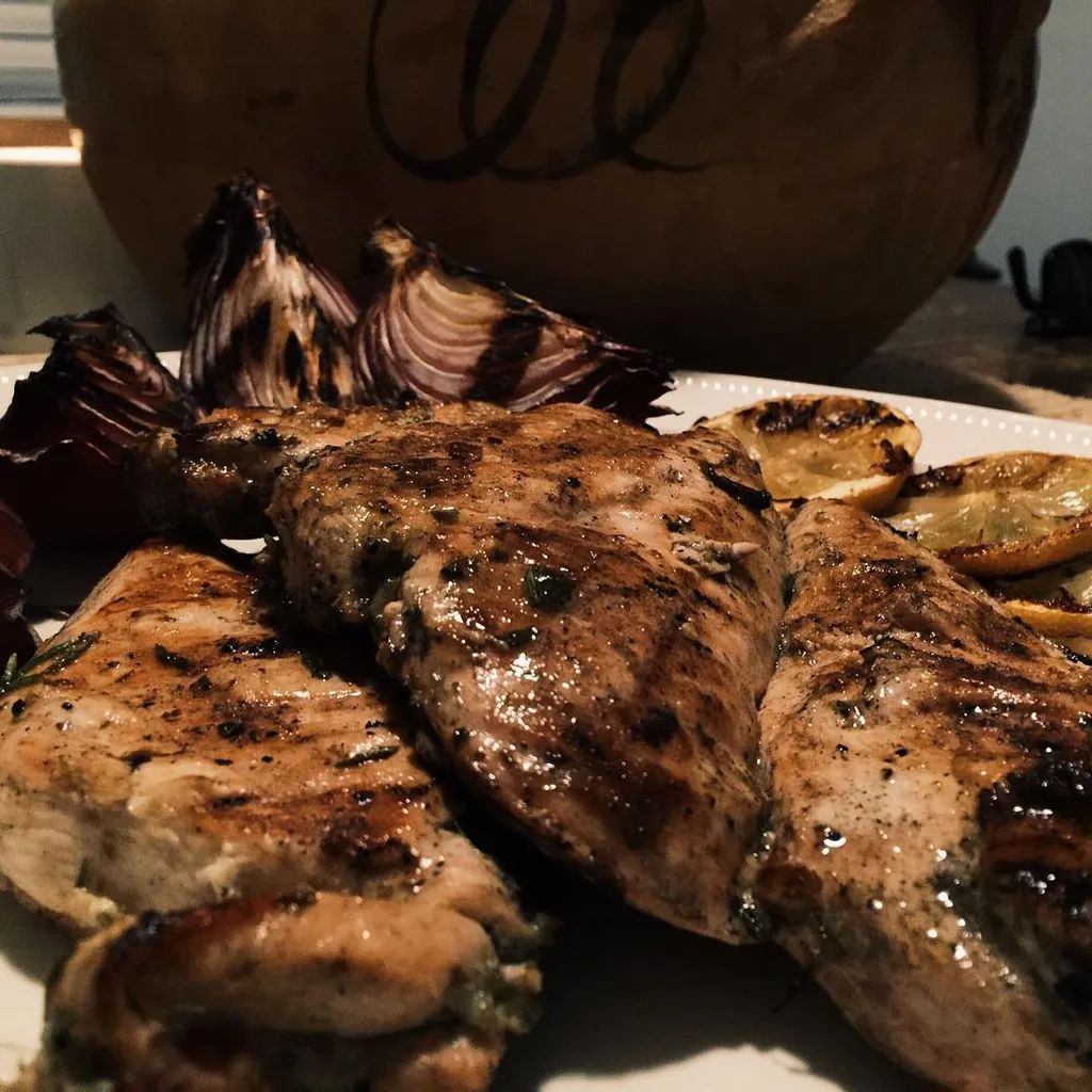 Grilled chicken breasts and roasted red onions are arranged on a white platter, set against a blurred background featuring a large, light brown, curved object.
