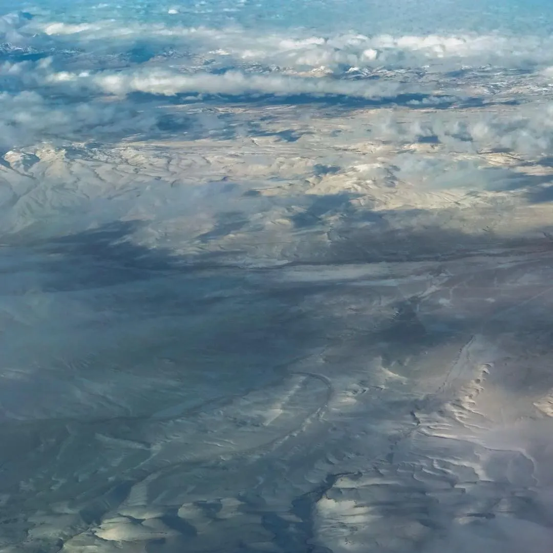 Horizontal composition of a muted gray and blue landscape surface, marked by intricate, swirling patterns and partially covered by scattered white cumulus clouds.