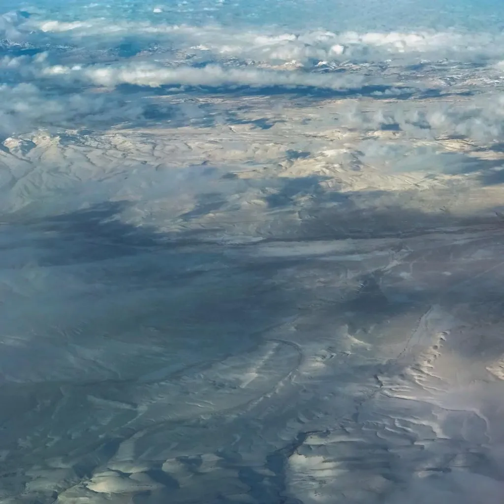 Horizontal composition of a muted gray and blue landscape surface, marked by intricate, swirling patterns and partially covered by scattered white cumulus clouds.