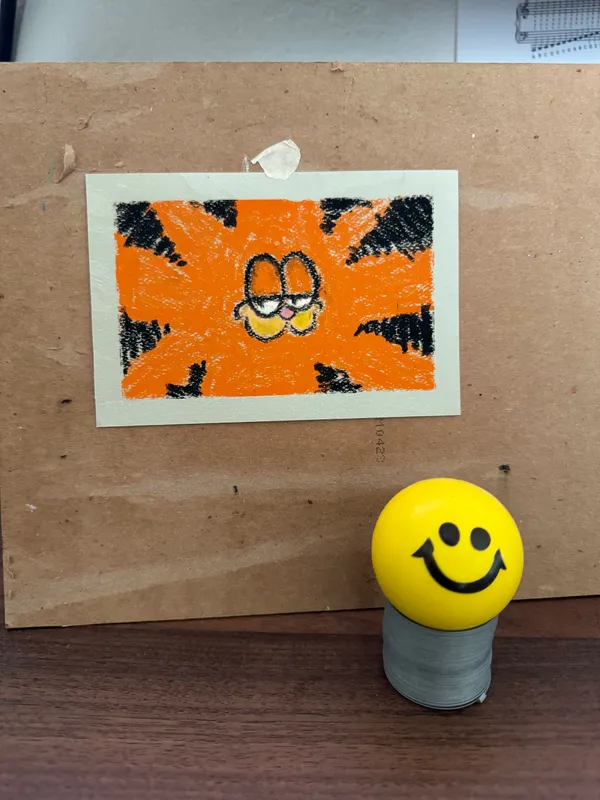 A square drawing featuring a yellow and black face against a bright orange background with radiating black marks is taped to brown cardboard, with a yellow smiley face on a gray cylinder resting below it.