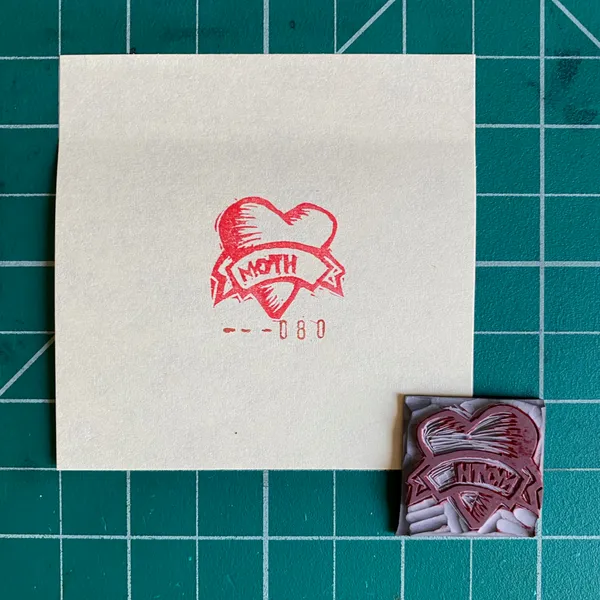 A cream card on a dark green grid background features a red ink design of a heart surrounding the word MOTH and the numbers --080, next to a smaller patterned red and white square.