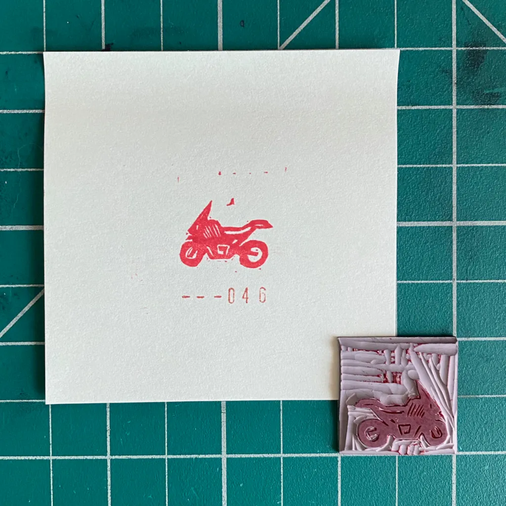A white card on a teal grid displays a red motorcycle graphic and the number --046, next to a small, textured square featuring an embossed red motorcycle.