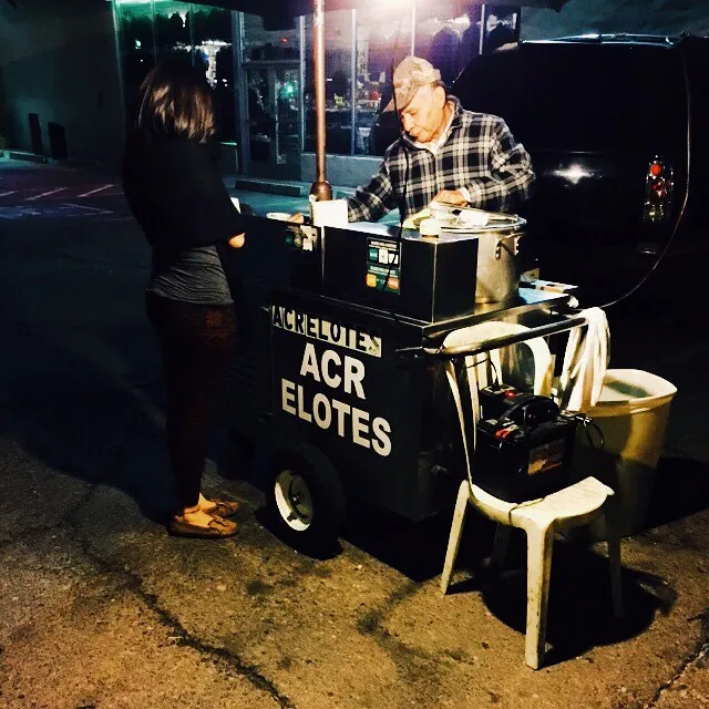 A dark street vendor cart with white lettering sits on concrete at night, flanked by a man operating the cart and a woman standing nearby under artificial lighting.