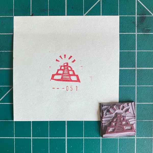 On a teal grid, a white card displays a red drawing of a stepped pyramid and the numbers --051, next to a small, textured, reddish-brown pyramid object.