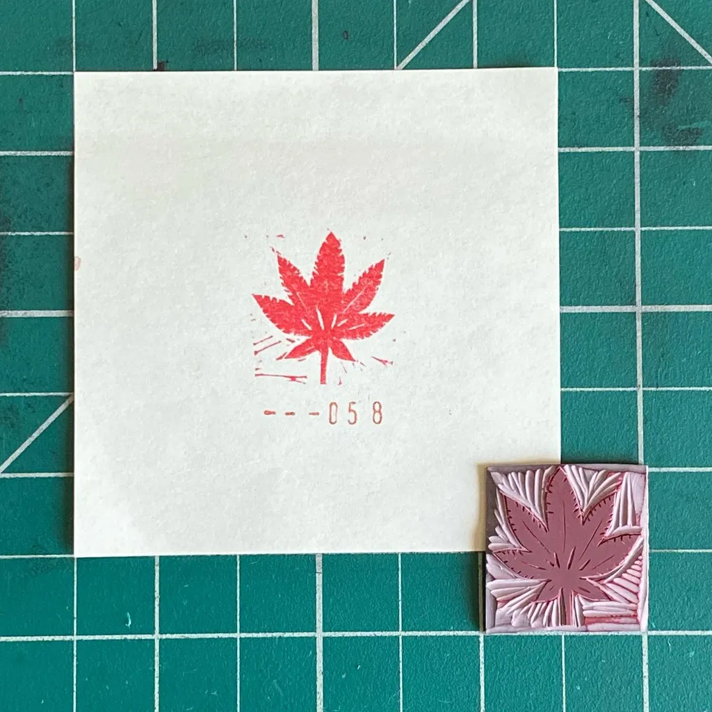 A white card displaying a red maple leaf and the number --058 rests on a green grid, alongside a small, layered square featuring a pink and white embossed maple leaf.