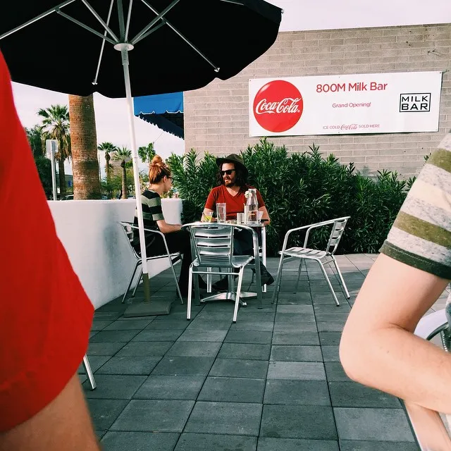 Under a large black umbrella, two people sit at small tables on a paved patio, with a cream building featuring a Coca-Cola sign and dense green foliage in the background.