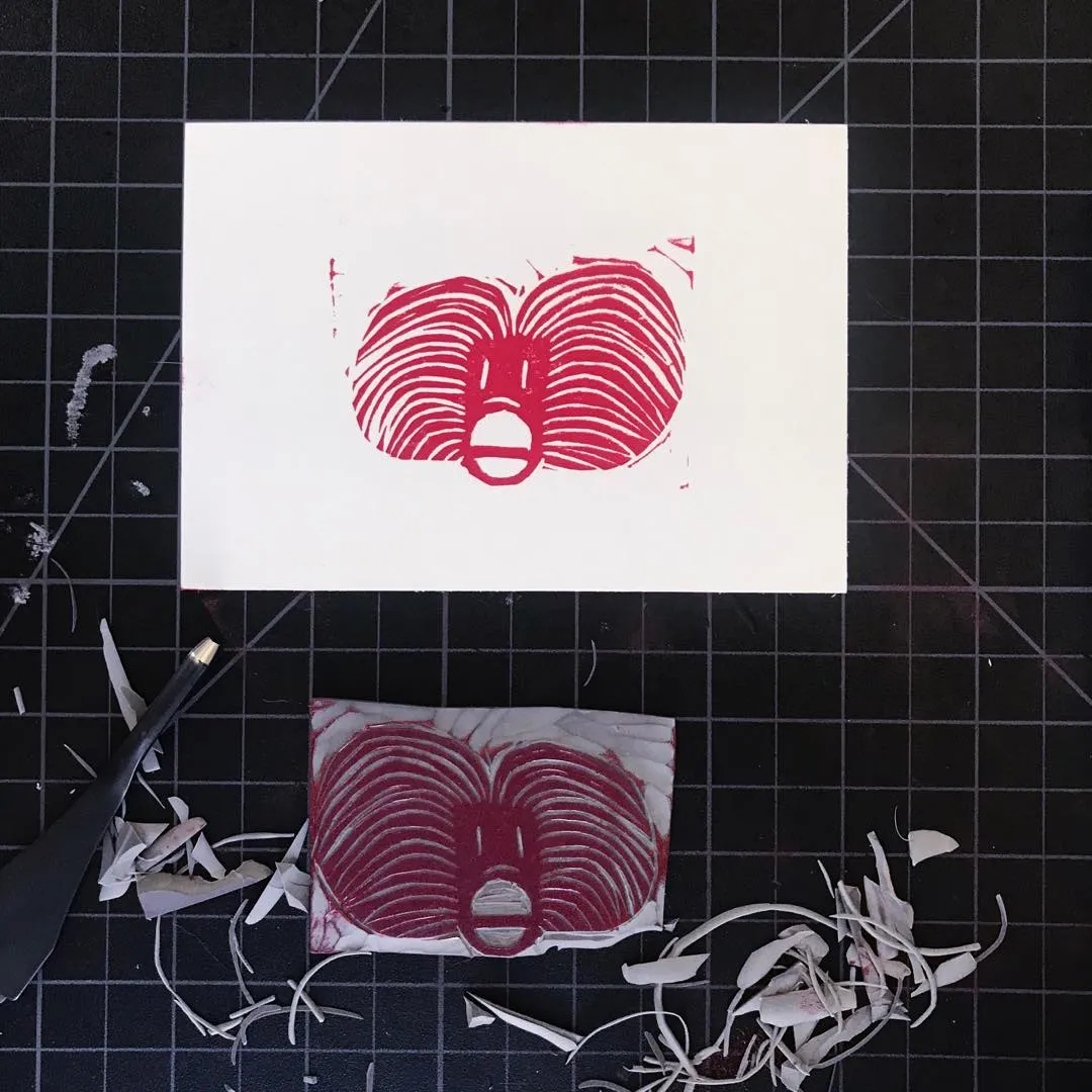 Three red drawings of a face, featuring wavy concentric lines, are displayed on white paper against a dark grid background, surrounded by scattered white and reddish-brown scraps.