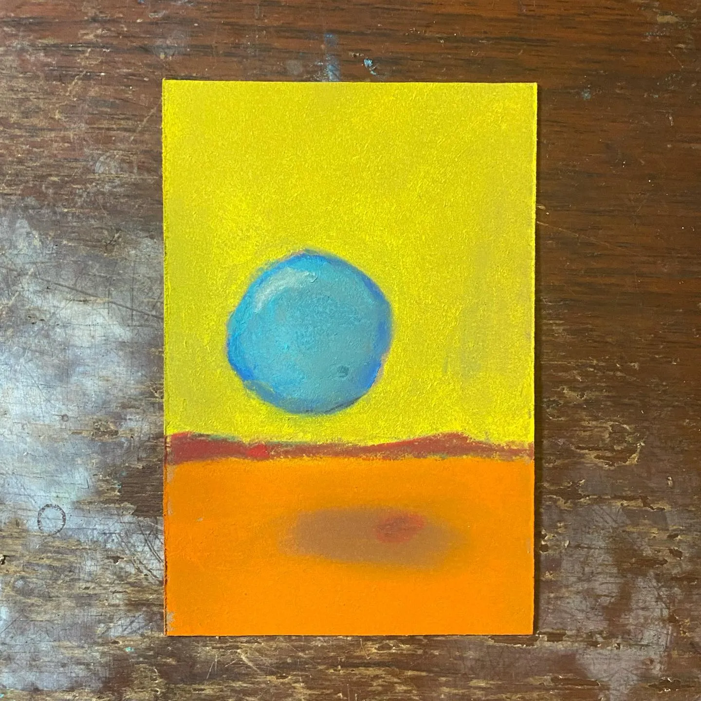 A square composition featuring a bright yellow upper field, a central blue circle, and a lower orange field with a darker, blended oval, set against a dark, distressed wooden background.