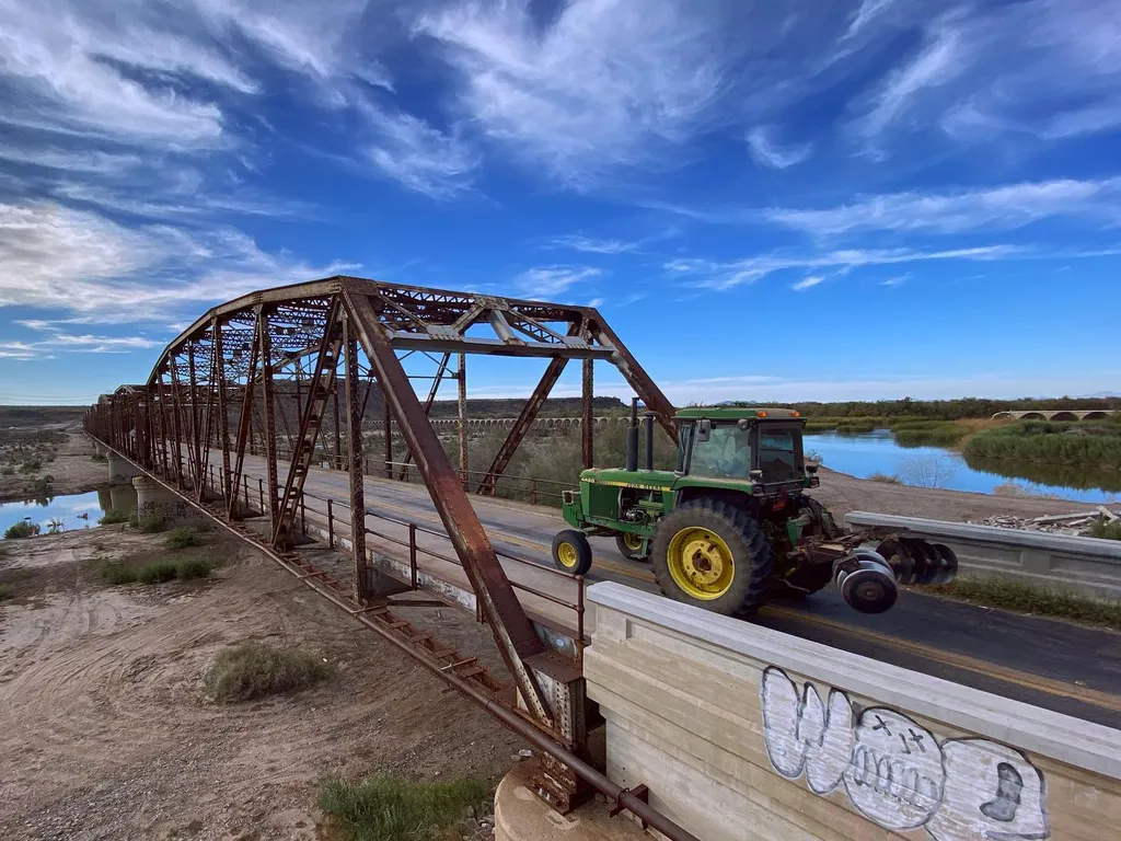 A rusty metal truss bridge spans a flat landscape under a blue sky, with a green truck driving across the road past a wooden barrier painted with white lettering.