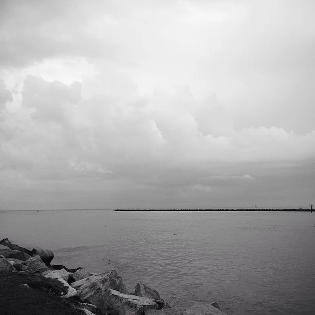 Rough rocks anchor the lower left corner, leading to a calm expanse of gray water beneath a vast, diffused sky of heavy clouds, with a low jetty visible on the distant horizon.