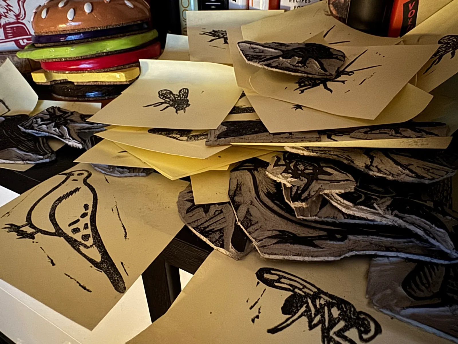 A stack of overlapping papers features various drawings of insects, including detailed black and white studies of wings and simpler sketches of flies, set against a background of muted cream and gray tones.