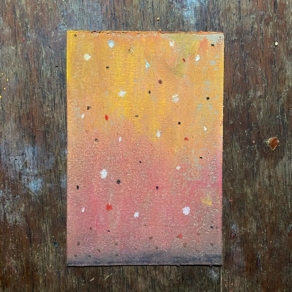 A rectangular artwork featuring a yellow-to-pink gradient, speckled with small white and colored dots, rests on a dark, weathered wooden surface.
