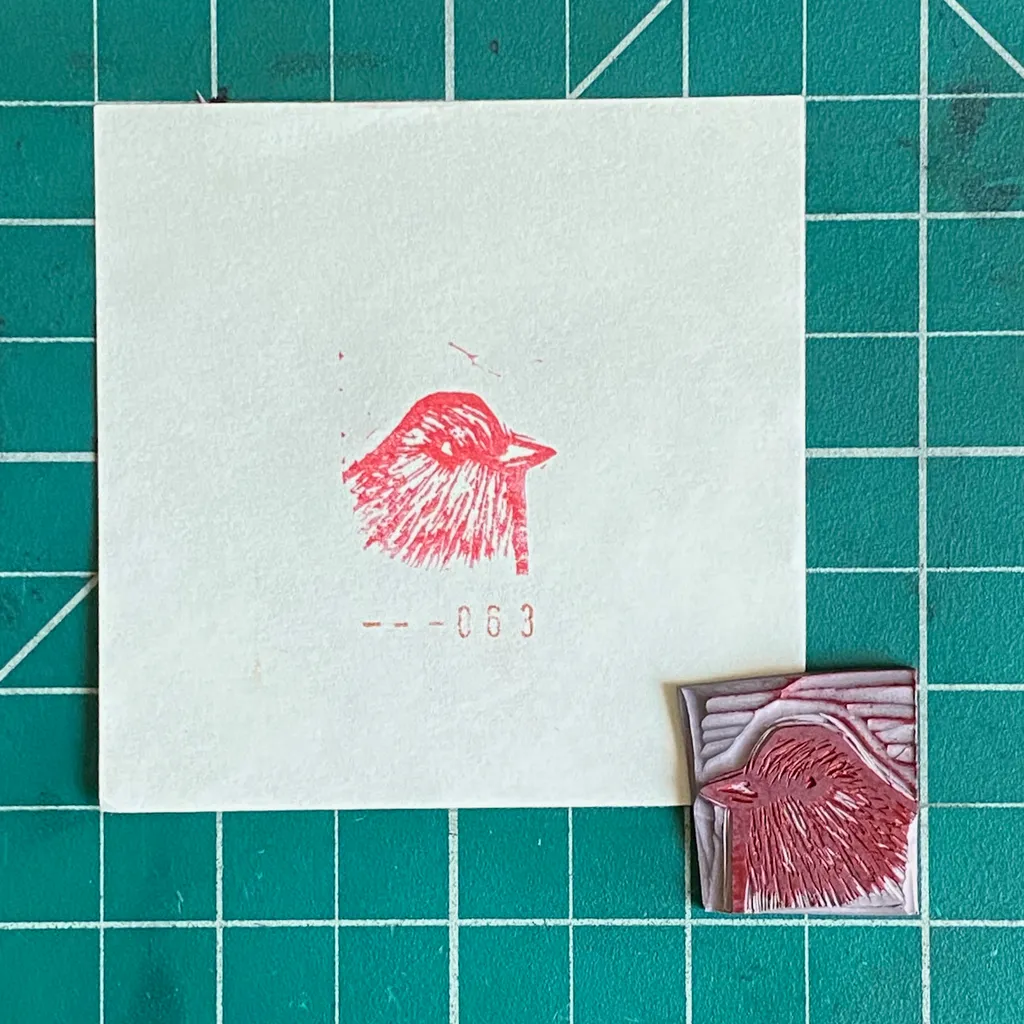 A white card on a teal grid displays a red, sketchy drawing of a bird head and the number -063, positioned next to a small square object featuring a textured, layered red bird form.