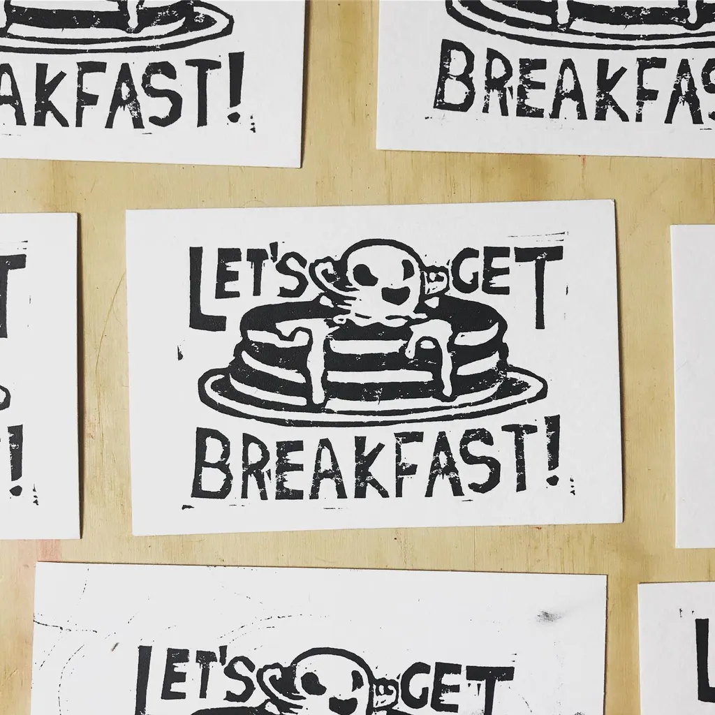 Multiple distressed black and white signs are arranged on a light wooden surface, featuring the phrase LET'S GET BREAKFAST! and an illustration of a skull sitting on a stack of pancakes.