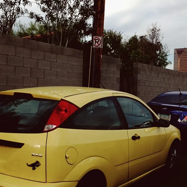 A bright yellow car is parked in the foreground before a tall, textured gray block wall, with green foliage visible above and a pole bearing a red and white sign standing nearby.