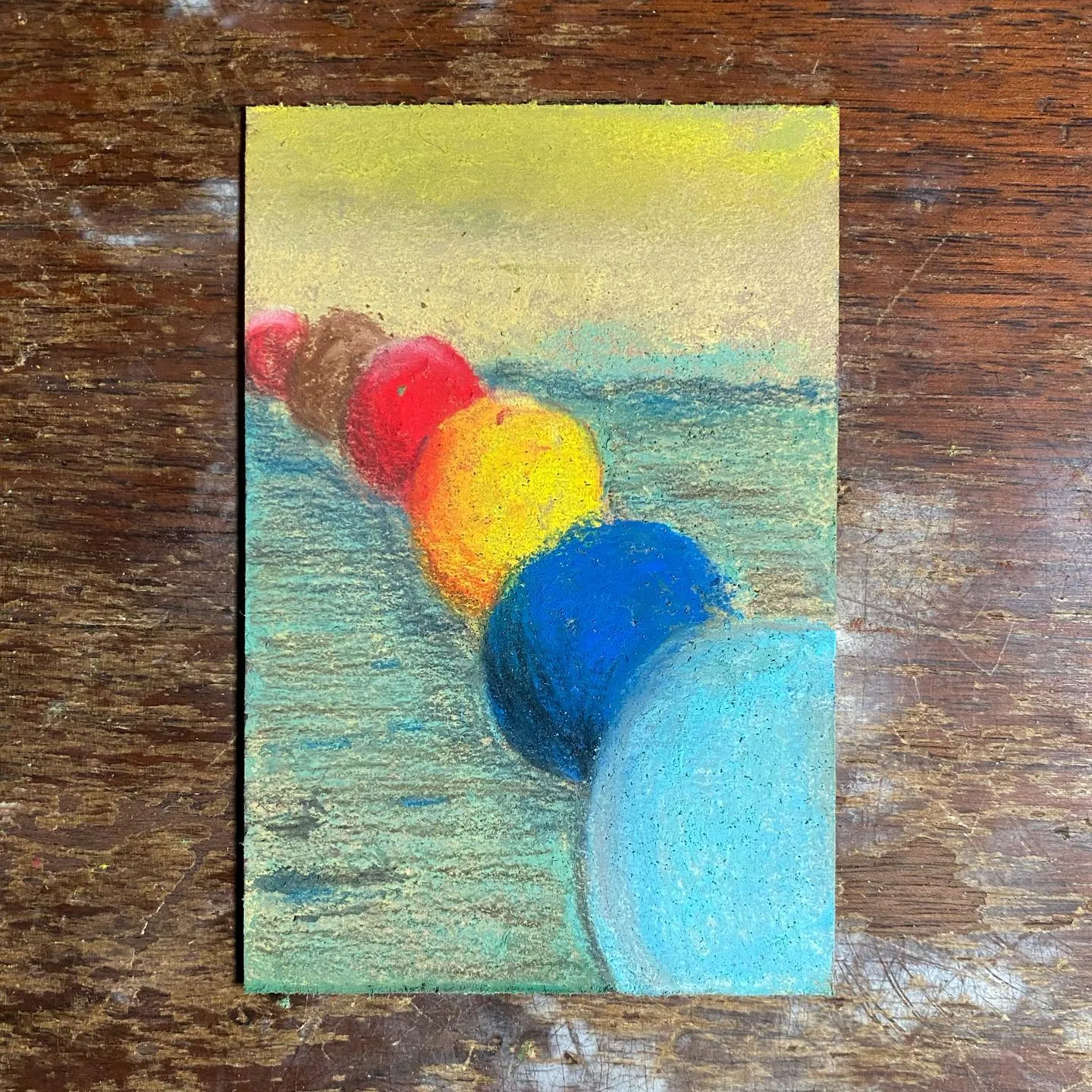 A horizontal row of brightly colored spheres—red, orange, yellow, dark blue, and light blue—are painted across a background divided into a pale yellow sky and textured greenish-blue water.