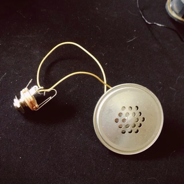 A small, silver mechanical assembly connected by a yellow wire to a circular, perforated metal disk rests on a dark, matte background.