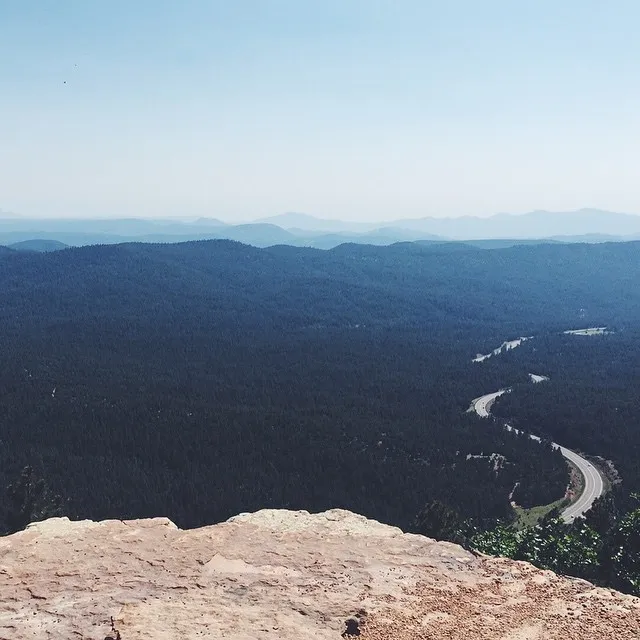 Rough tan rock occupies the foreground, overlooking a vast expanse of deep green forest, a winding road, and receding layers of hazy blue mountains beneath a clear sky.