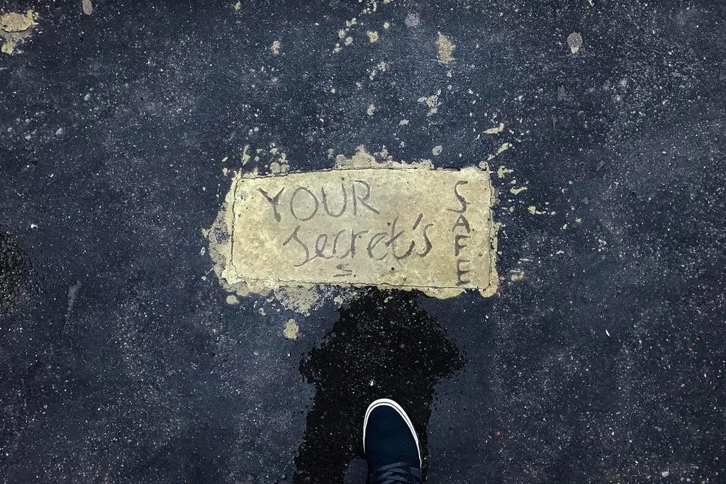 Dark, speckled ground fills the frame, featuring a light rectangular patch with the handwritten phrase YOUR SECRETS SAFE, centered above a pair of shoes.