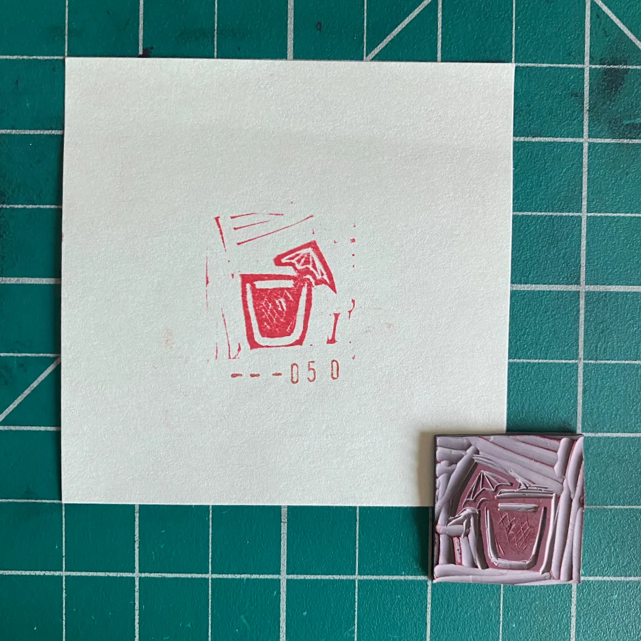 On a teal grid, a white paper displays a red sketch of a drink and the code -050, next to a small, rectangular, layered object featuring abstract pink and purple tones.