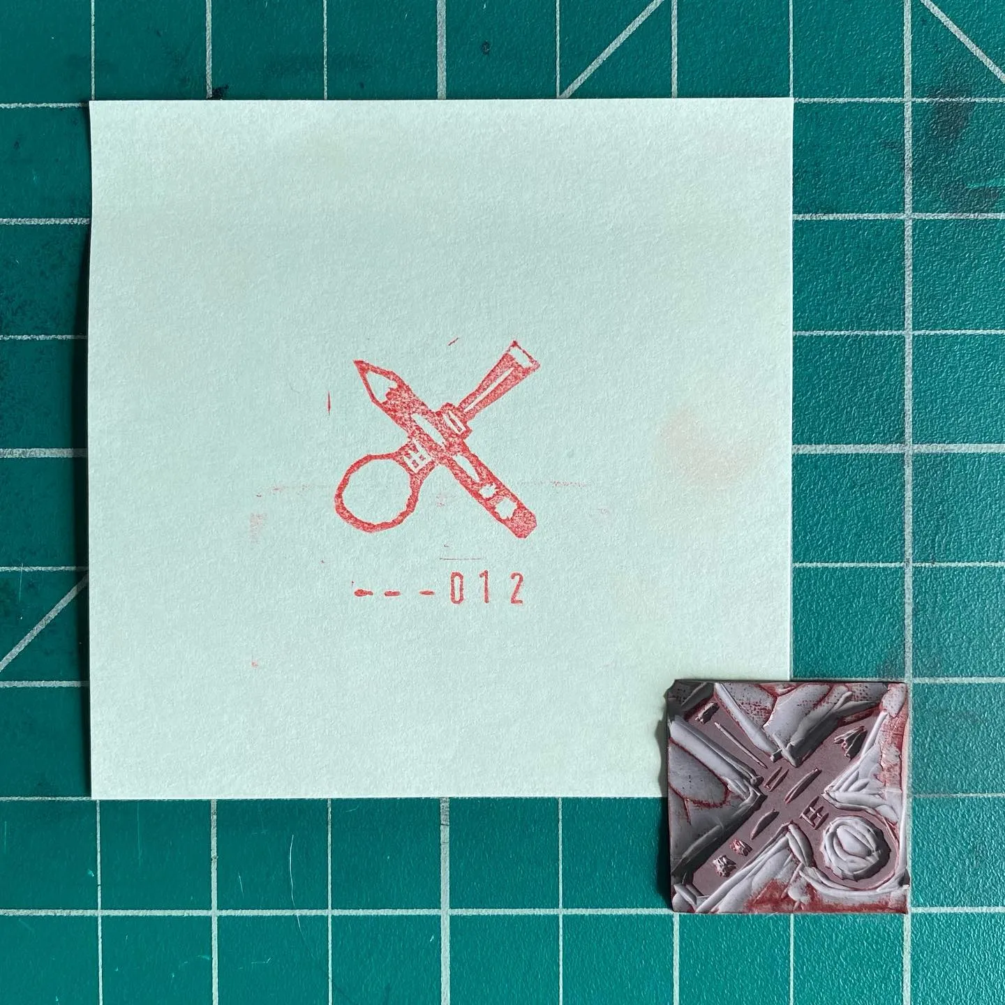 A light square paper on a teal grid features a red outline drawing of crossed scissors and the text -- 012, next to a small, textured square object with abstract red and white shapes.