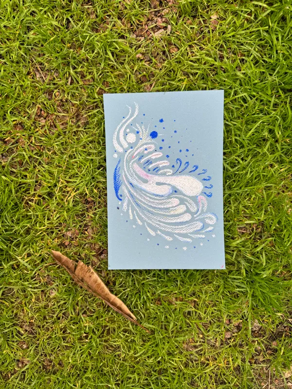 On vibrant green grass, a light blue card displays a flowing white and blue feather design with scattered blue dots, flanked by two small, brown, dried plant pieces.