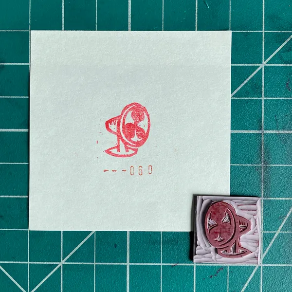 A white card with a red ink drawing of a stylized face and the numbers ---060 rests on a green grid background, next to a small square object featuring a similar red face design.