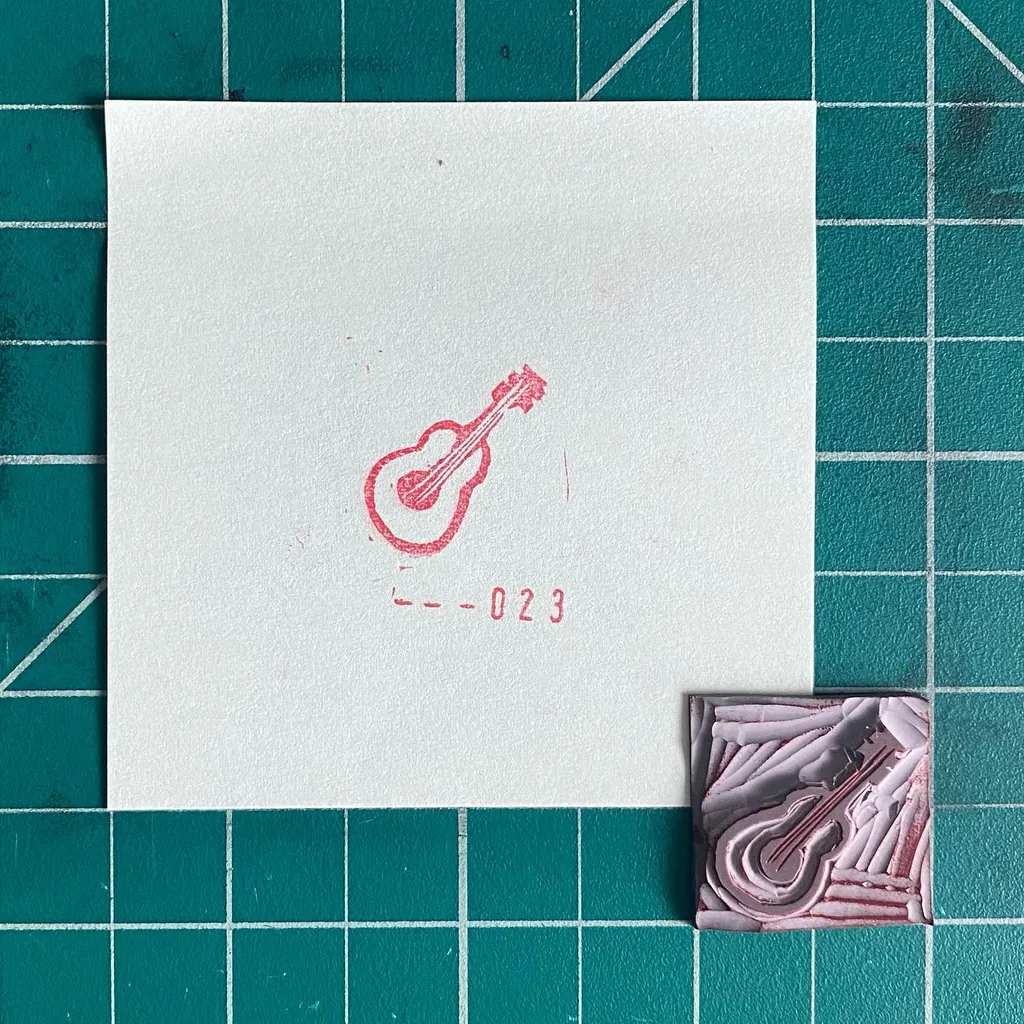 The composition features a white card on a teal grid background, displaying a red outline drawing of a guitar and the number -023, alongside a small, textured square piece in purple and white.