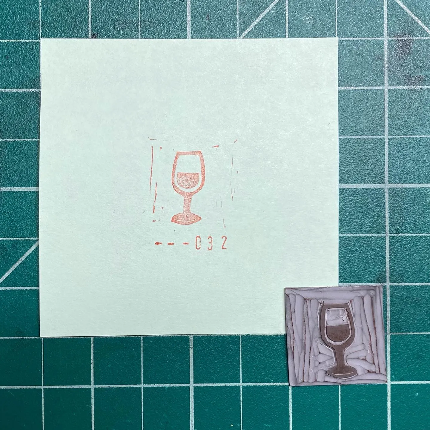 A light square paper on a teal grid displays a red pencil drawing of a wine glass and the code --032, positioned above a small, square, textured piece featuring a sculpted wine glass.