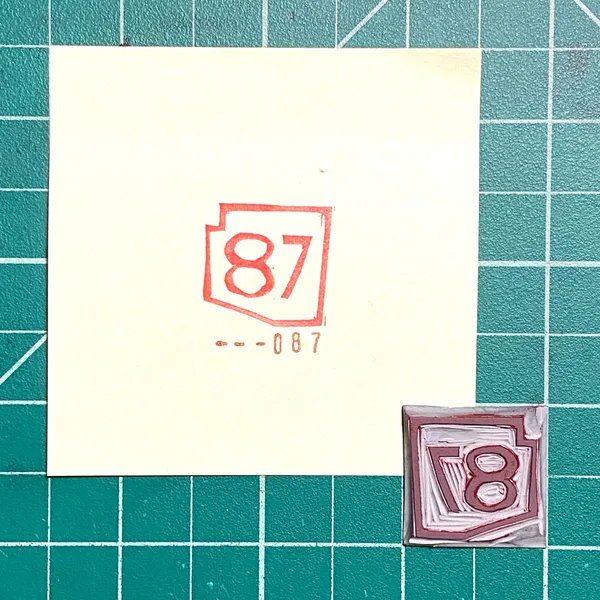 A cream paper on a teal grid shows red outlines of 87 and --087, next to a small square painted with a stylized brown number 8.