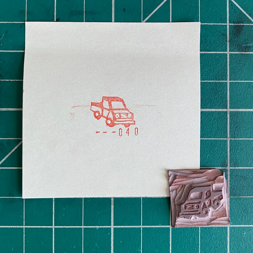 On a green grid, a white paper displays a red outline drawing of a car and the numbers --040, next to two small, textured pieces, one featuring carved Arabic script.