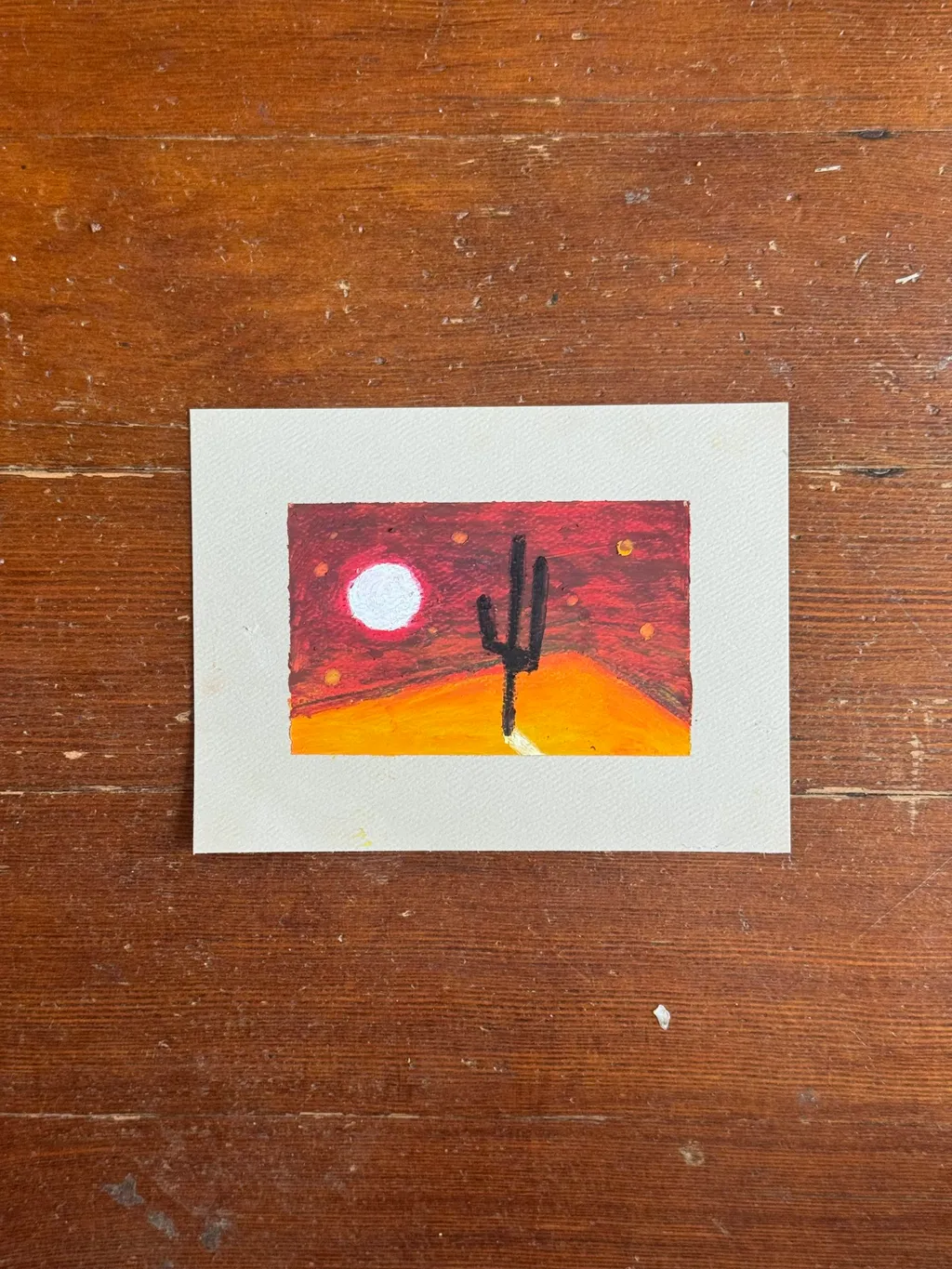 A small, square painting depicts a desert scene with a black saguaro cactus silhouetted against a yellow-orange ground and a deep red sky featuring a white circle.