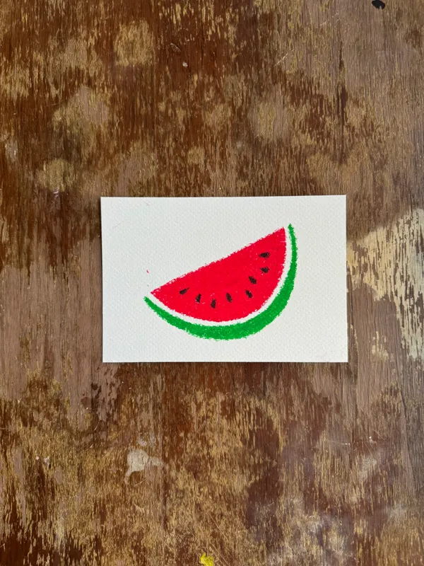 A drawing of a red and green watermelon slice sits on a white paper square, which is set against a richly textured, dark brown wooden background.