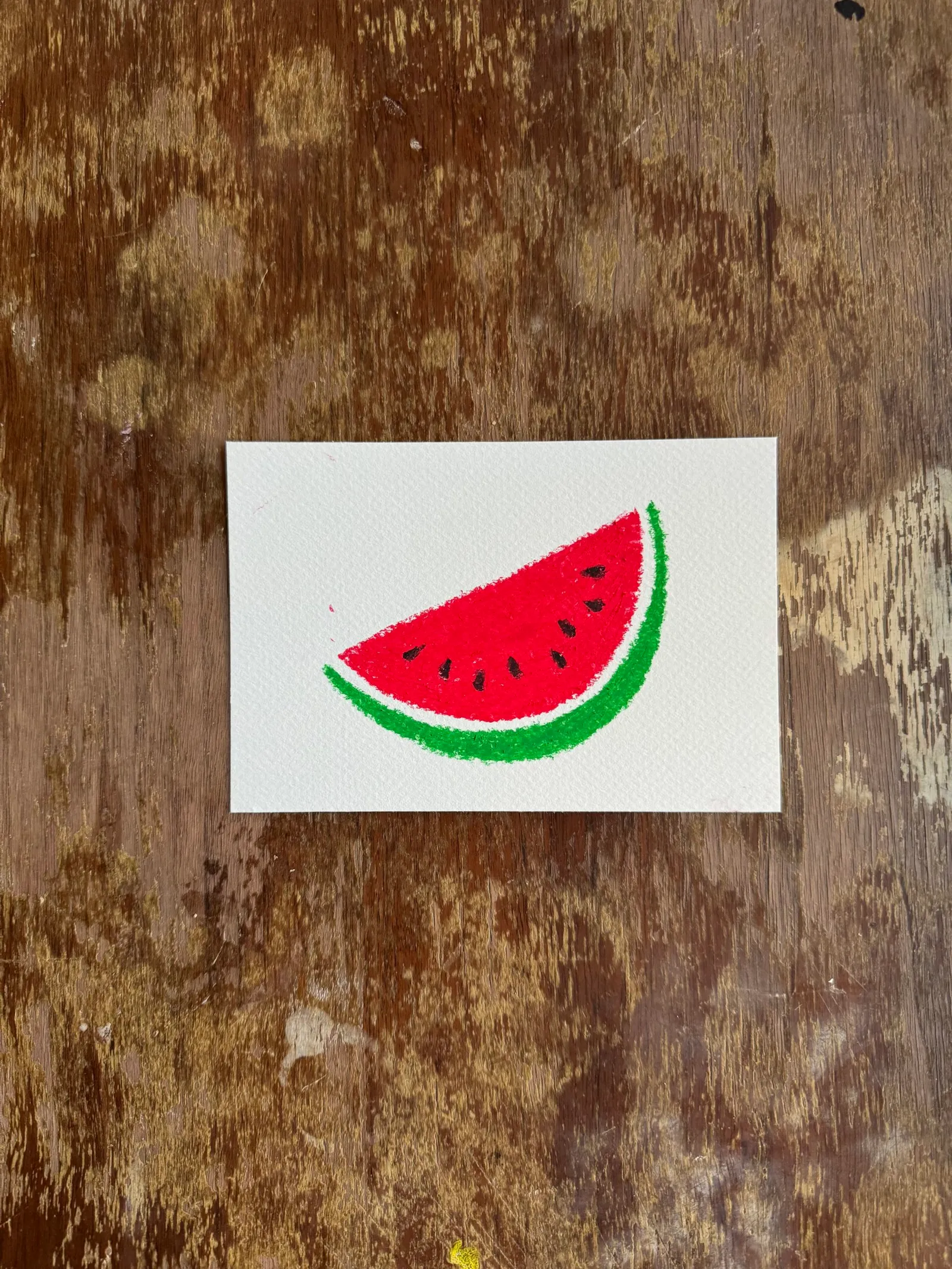 A drawing of a red and green watermelon slice sits on a white paper square, which is set against a richly textured, dark brown wooden background.