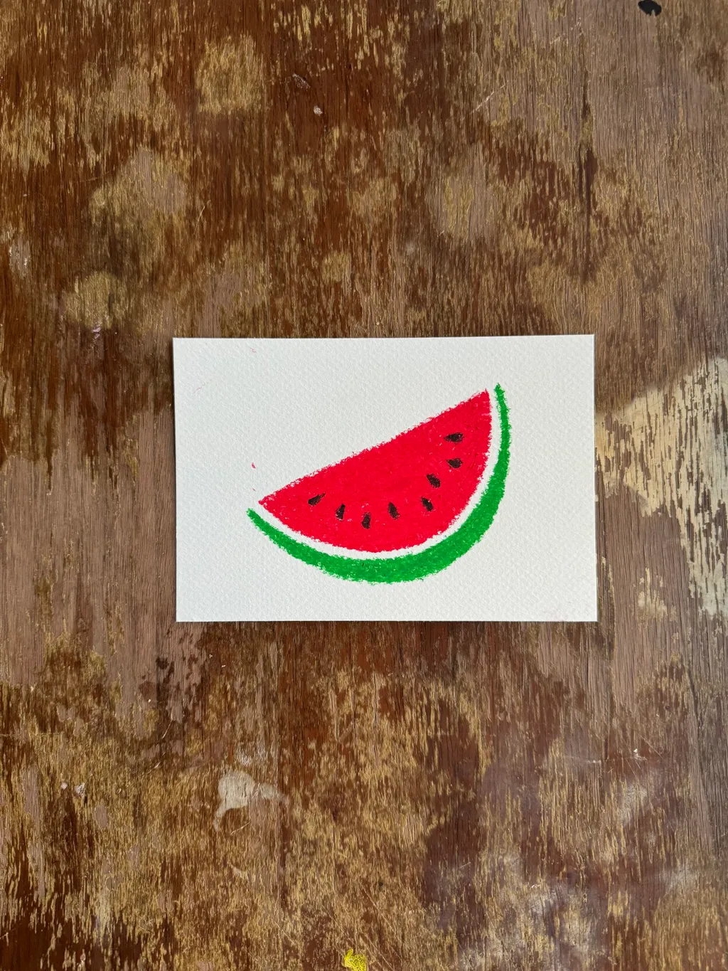 A drawing of a red and green watermelon slice sits on a white paper square, which is set against a richly textured, dark brown wooden background.