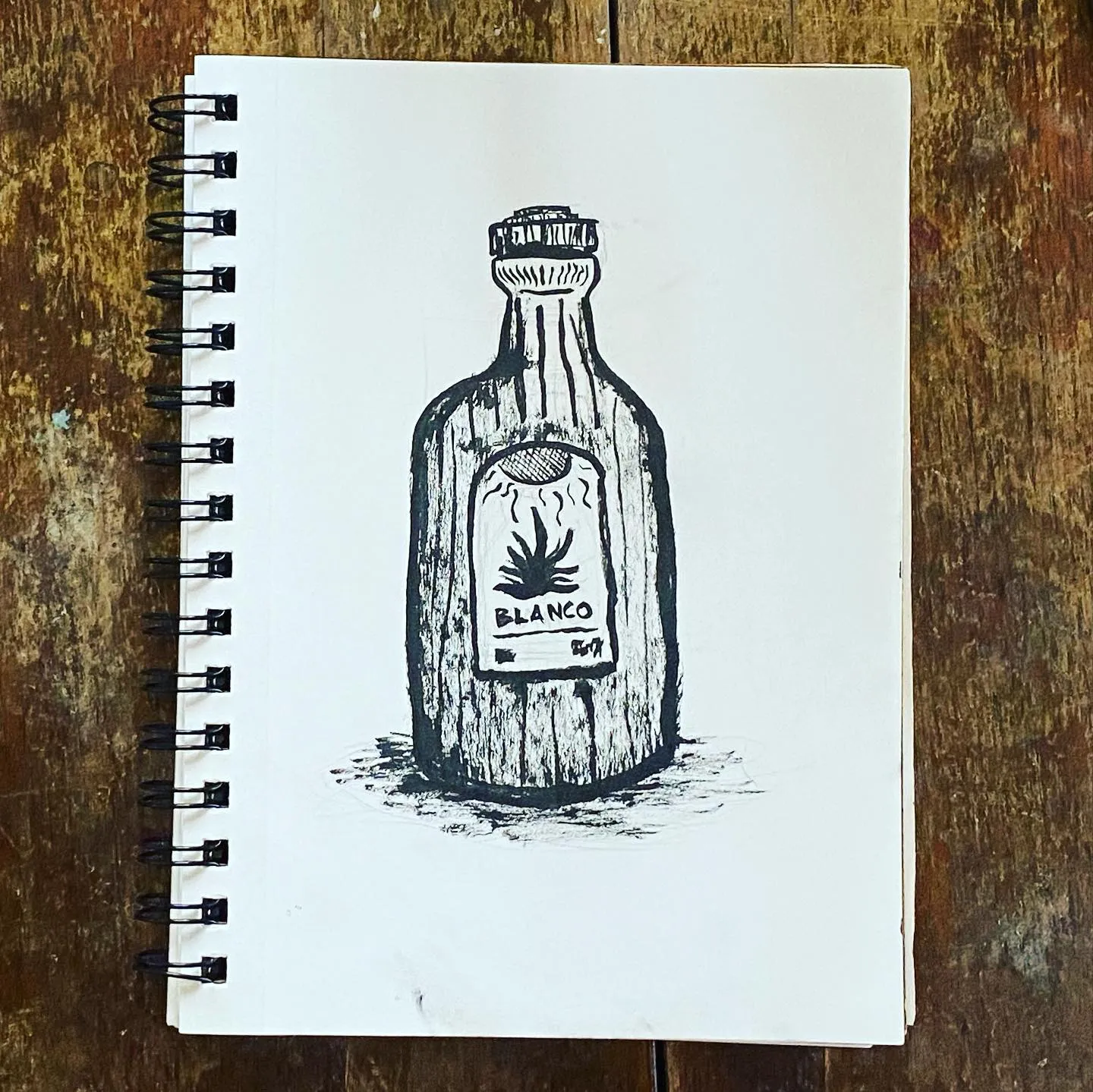 A black ink drawing of a stylized liquor bottle on a white sketchbook page, set against a dark wooden background, featuring sketchy lines and a label with a plant drawing.