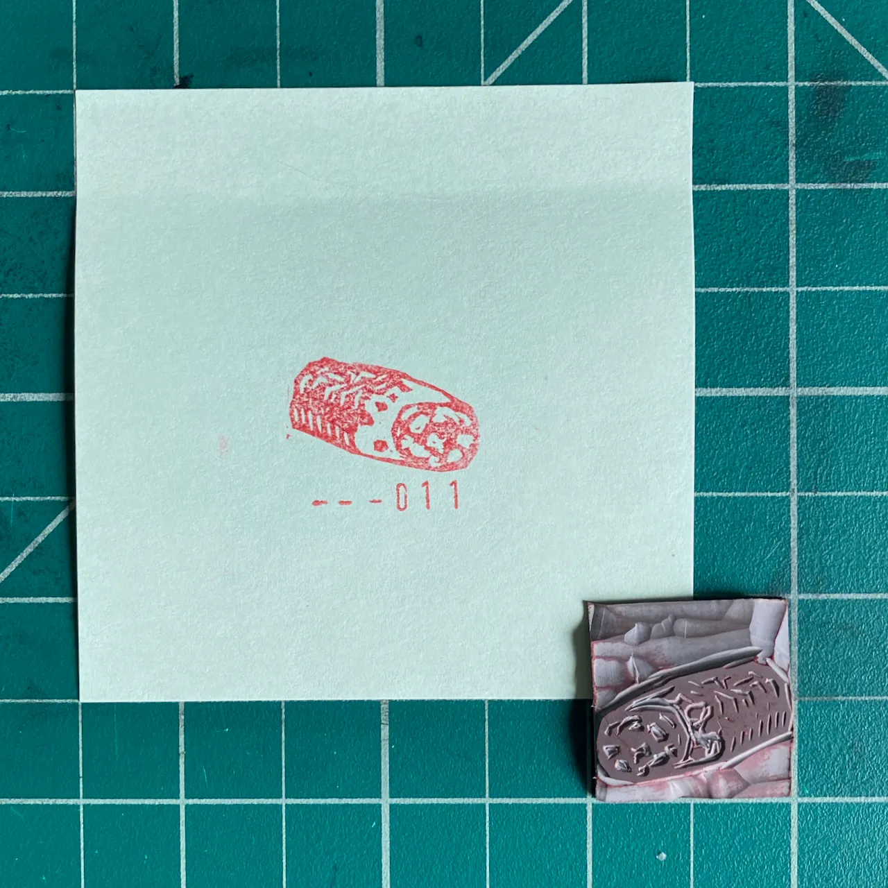 On a teal grid, a mint square paper displays a red drawing of an oval object and the code -011, accompanied by a small, textured, pinkish-mauve piece.