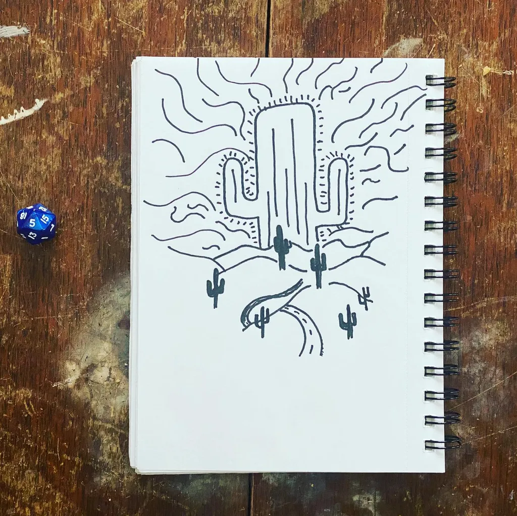 A black line drawing of a large saguaro cactus in a desert landscape, resting on an open sketchbook on a dark, textured wooden surface, with two blue dice nearby.