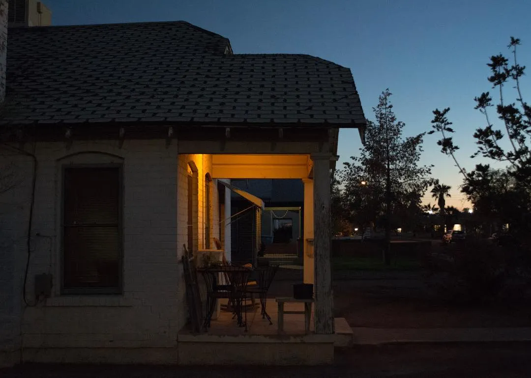 A single-story building with a tiled roof is viewed at dusk, featuring a warm yellow glow emanating from its open doorway, set against cool blue and gray tones.
