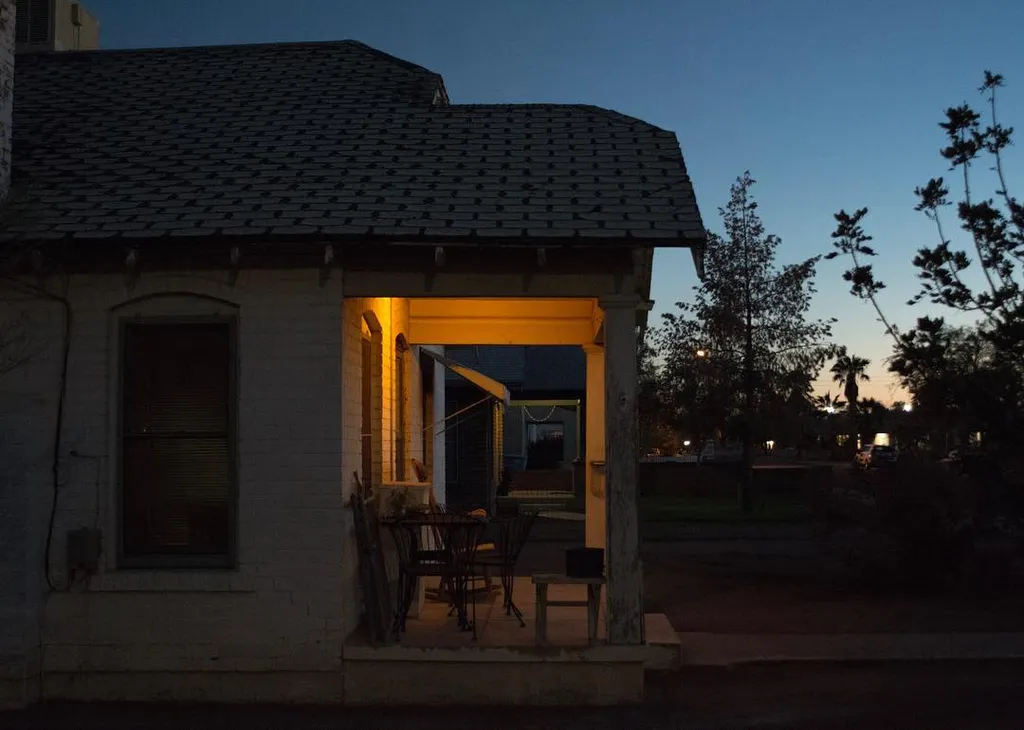 A single-story building with a tiled roof is viewed at dusk, featuring a warm yellow glow emanating from its open doorway, set against cool blue and gray tones.